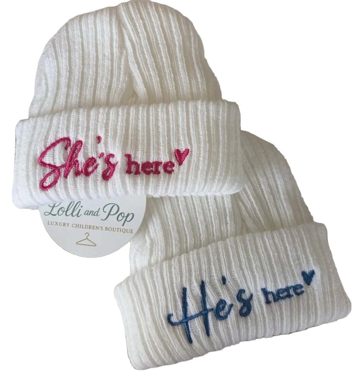 White ribbed knit baby beanie hats on a striped surface. One hat has pink embroidered text reading 'She's here' with a small pink heart, and the other has blue embroidered text reading 'He's here' with a small blue heart.